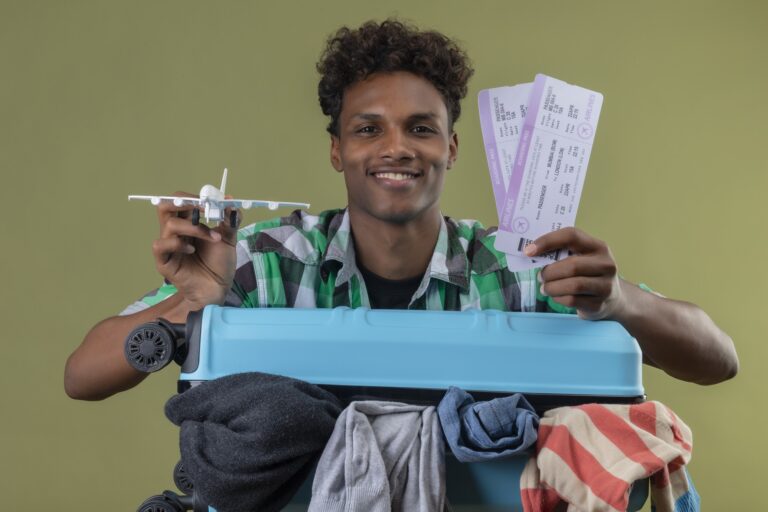 young-african-american-traveler-man-standing-with-suitcase-full-clothes-holding-air-tickets-toy-air-plane-looking-camera-smiling-happy-positive-green-background