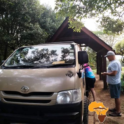 A person enters a tan van parked near a wooden structure, surrounded by trees and greenery. Another person stands nearby. A logo of a stylized African continent with orange and brown colors is visible on the bottom right.