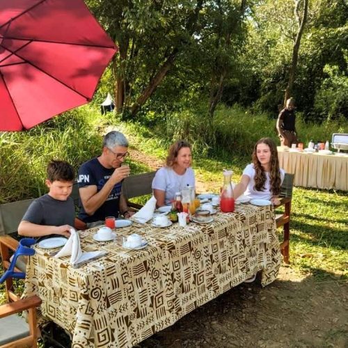 A family of four sits outdoors at a table covered with a patterned cloth. They are enjoying breakfast with various dishes and drinks. A large red umbrella provides shade, and trees and a buffet table are visible in the background.